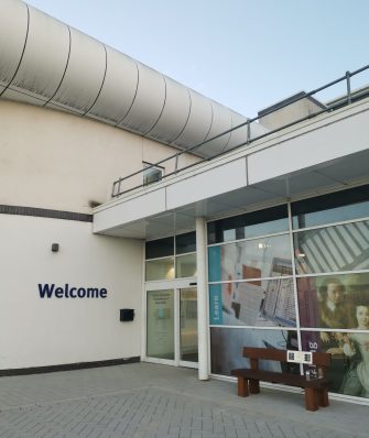 Photograph of the front entrance of the Essex Record Office, a large white building with 'Welcome' in blue letters on the side and a wooden bench to the front.